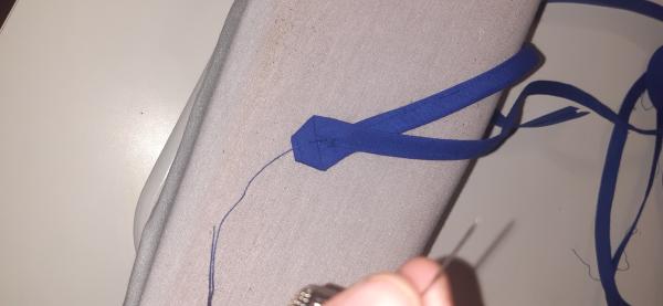 Ici, je suis en train de confectionner un des 3 motifs qui iront sur le devant de la veste. Il s'agit du biais bleu préparé précédemment, entrelacé, et cousu dans cette position à la main afin qu'il ne se déplie pas.<br>Normalement, il était prévu pour cette veste qu'elle aie un biais bleu sur tous ses bords. Mais en raison de l'épaisseur de mon tissu, je me suis rendu compte que cela serait difficilement possible.<br>Avec l'aide de mon père, j'ai trouvé l'idée de confectionner des motifs avec le biais et de les coudre sur le devant de la veste, pour remplacer les liserés manquants sur les bordures.